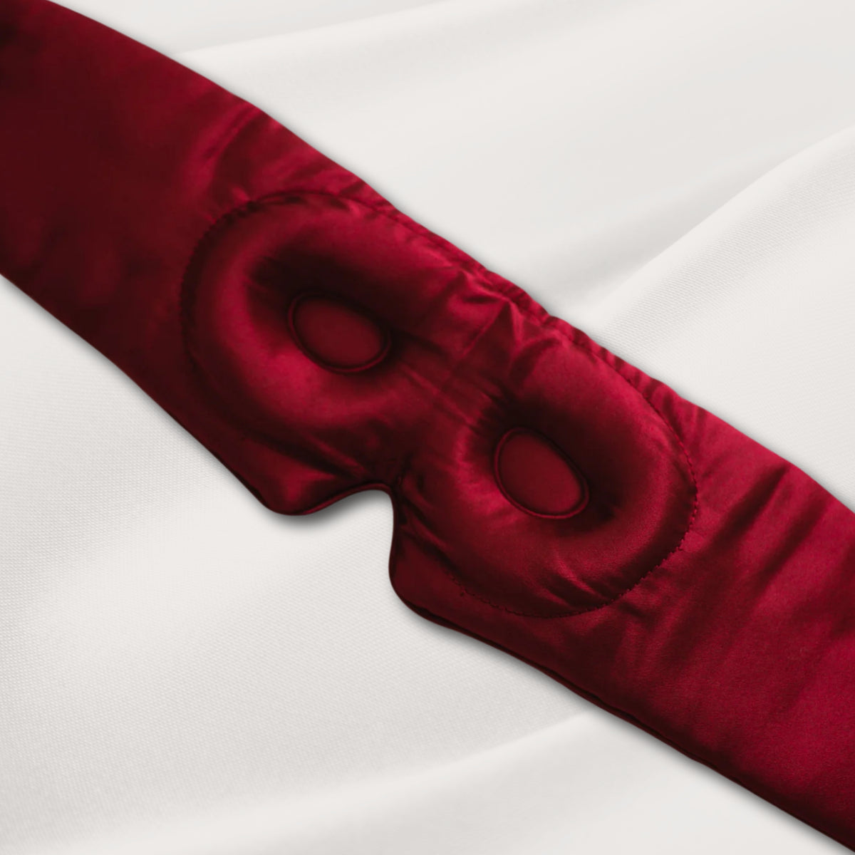 Burgundy LashGuard 3D sleep mask resting on white bedding, angled view of the contoured eye cups